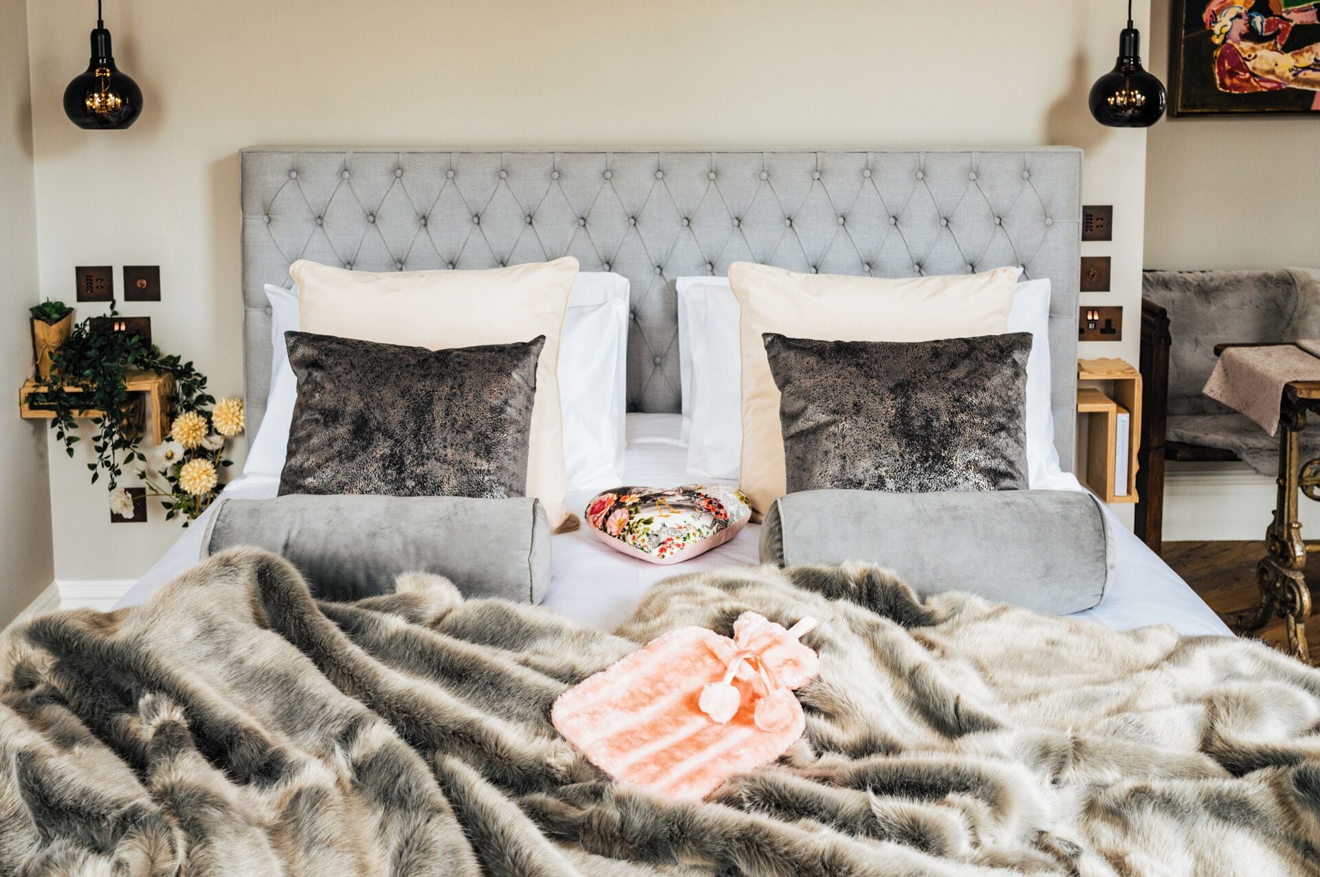 A bedroom with a tufted headboard, two white pillows, two dark pillows, a floral throw, and a pink towel.