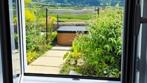 A view from a room with a glass door, overlooking a garden with a hot tub, a stone wall, and a mountain landscape. - Benilde Village House (Povoacao)