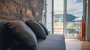 A modern room with a grey sofa, a stone wall, and a view of a mountain through large glass doors. - Benilde Village House (Povoacao)