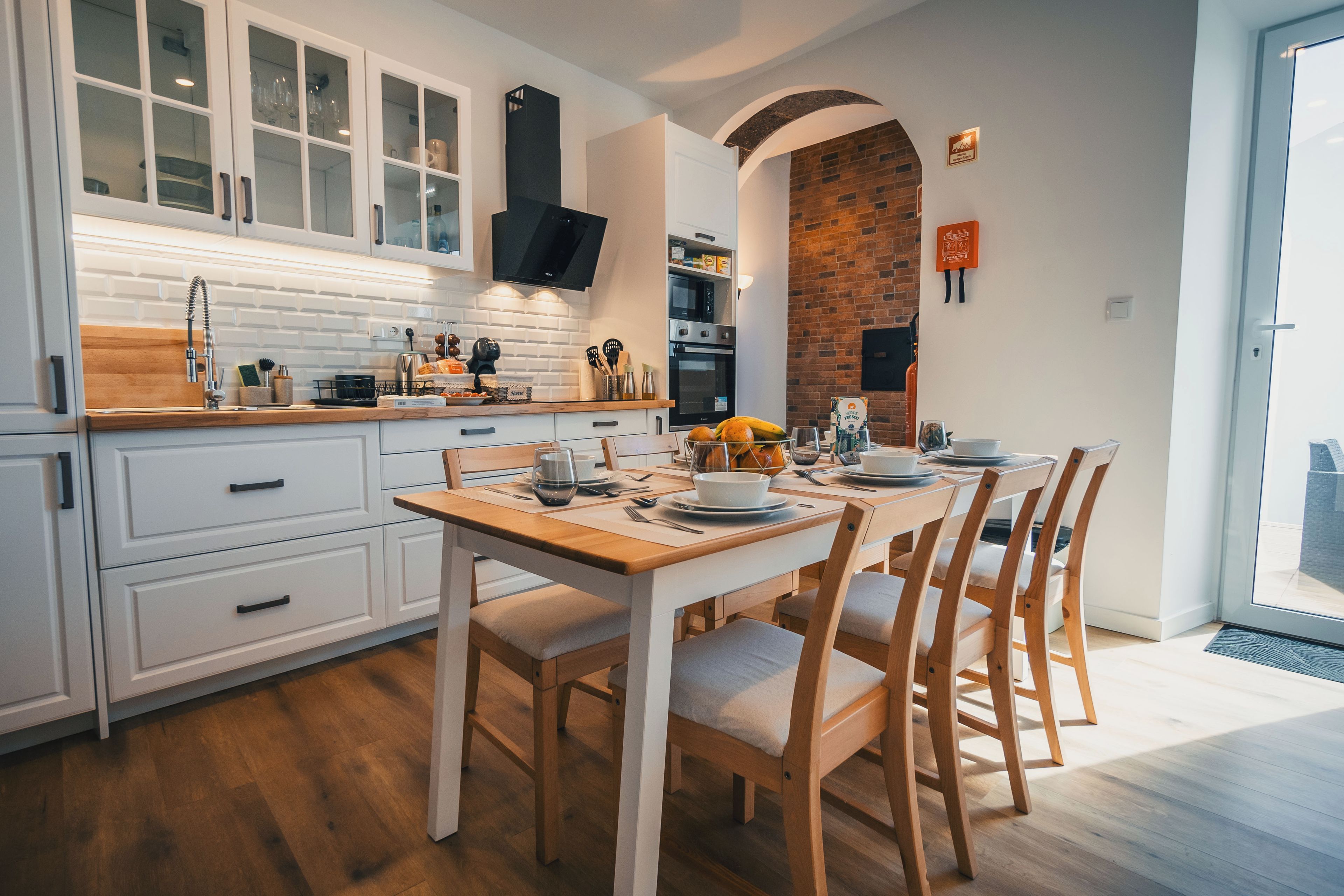 Una cocina moderna con muebles blancos, una mesa de comedor de madera y una pared de ladrillo.