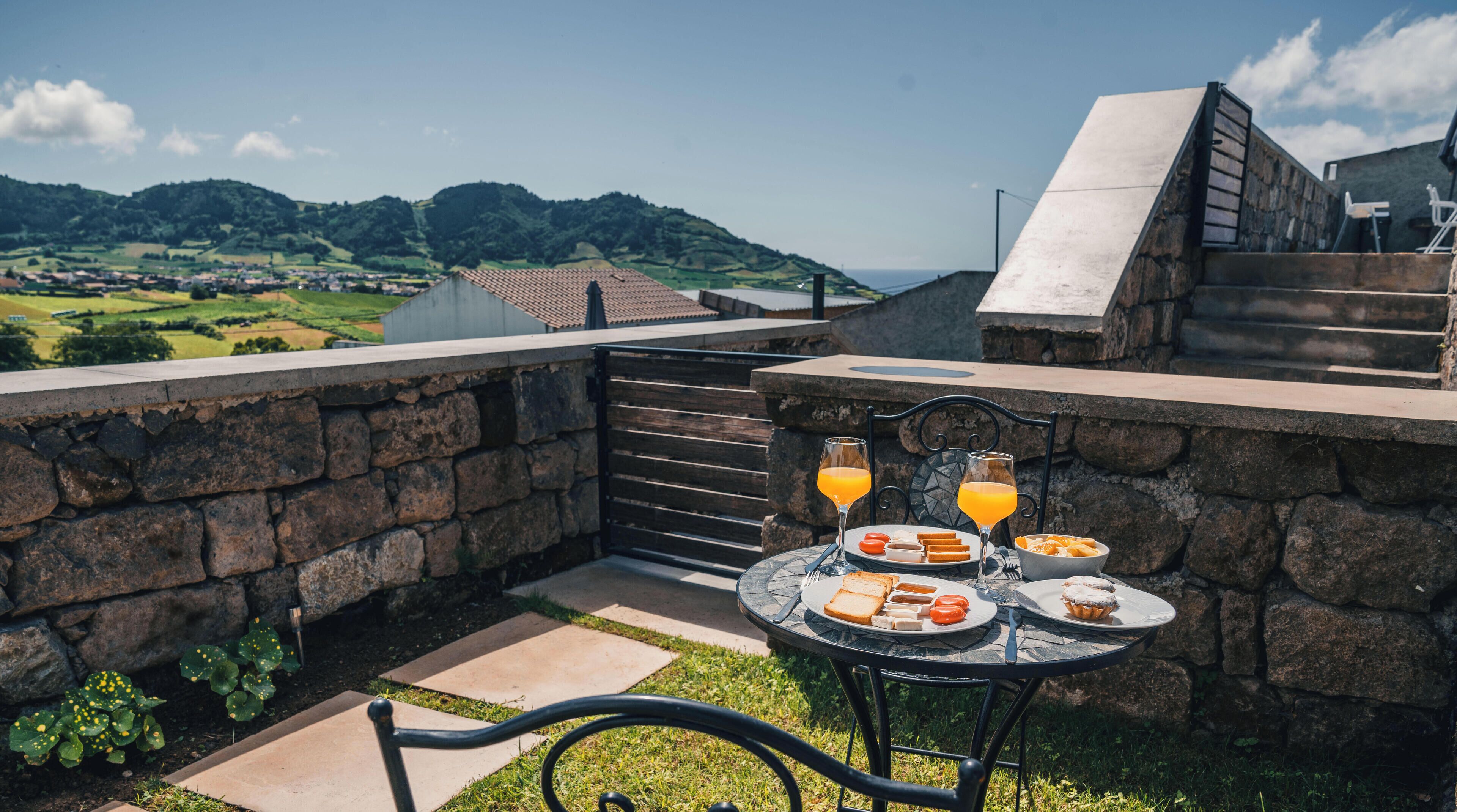 Un mur de pierre avec une table dressée pour le petit-déjeuner, comprenant des toasts, des œufs et du jus d’orange, donnant sur un paysage pittoresque avec des collines et des bâtiments.