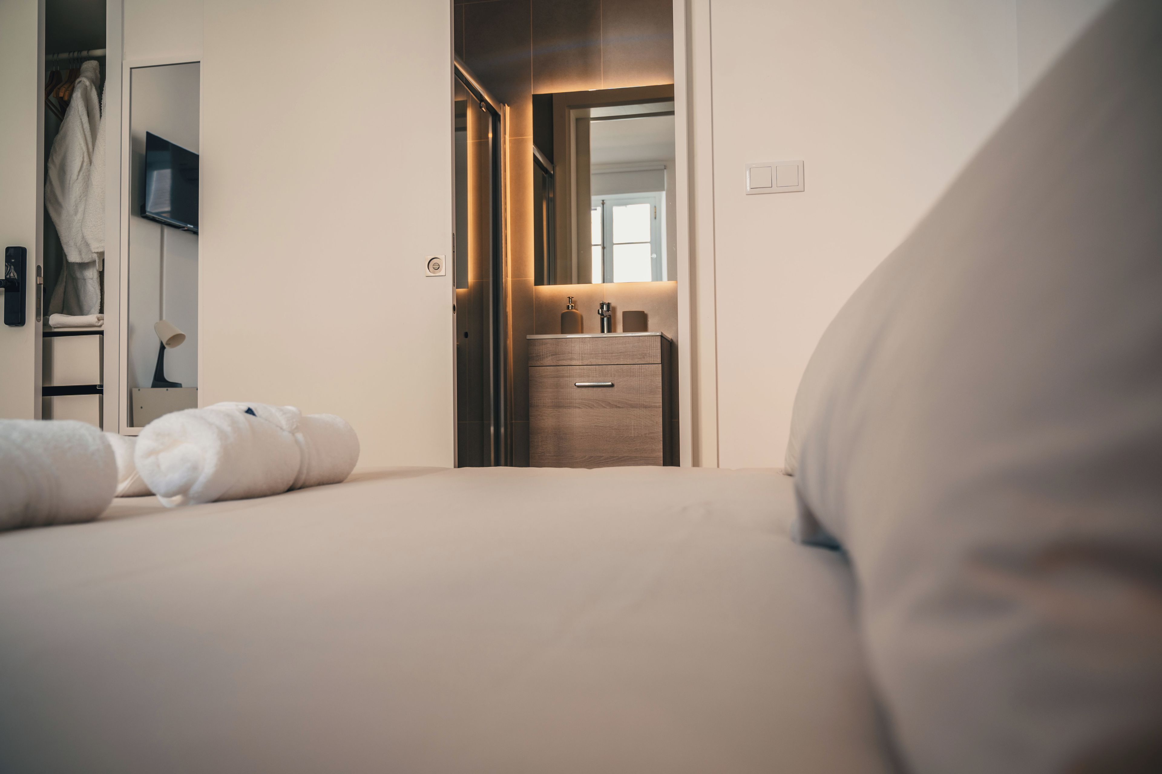 Habitación de hotel con cama, mesita de noche, espejo y baño con lavamanos y toallero.