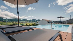 Un espace aménagé au bord de la piscine, avec des chaises longues, une piscine et une vue sur un village et des collines en arrière-plan.