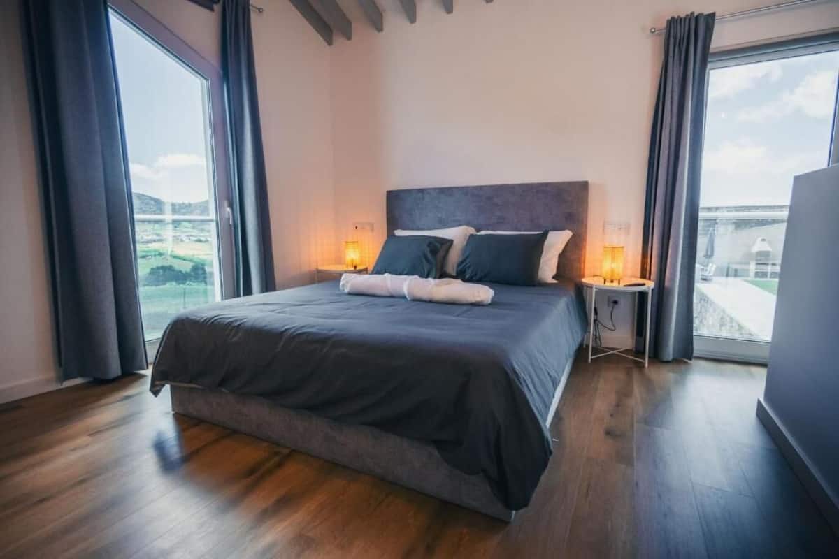Ein modernes Schlafzimmer mit einem großen Bett, zwei Nachttischen mit Lampen und Blick ins Freie durch große Fenster.
