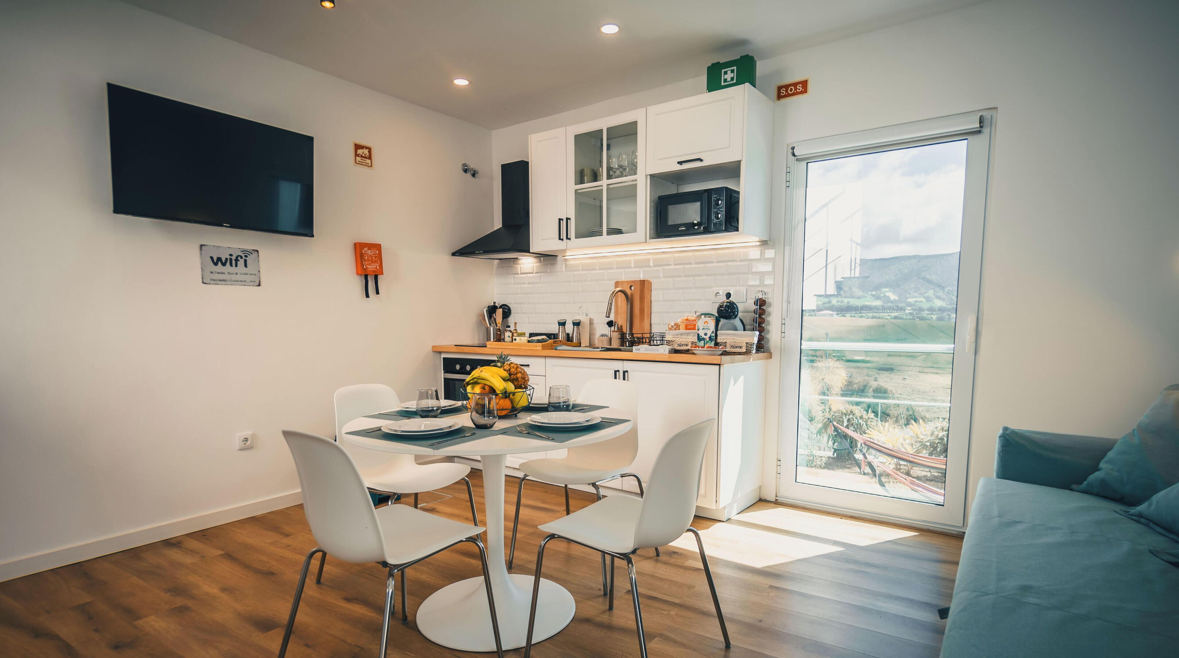 A modern kitchen with white cabinets, a microwave, and a sink. There is a dining table with chairs and a small fruit bowl. A TV is mounted on the wall, and a window provides a view of the outdoors.