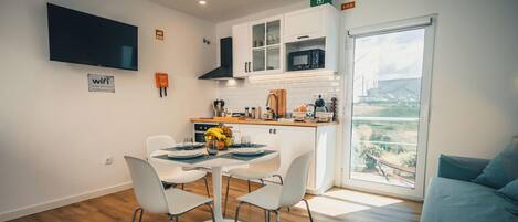 A modern kitchen with white cabinets, a microwave, and a sink. There is a dining table with chairs and a small fruit bowl. A TV is mounted on the wall, and a window provides a view of the outdoors.