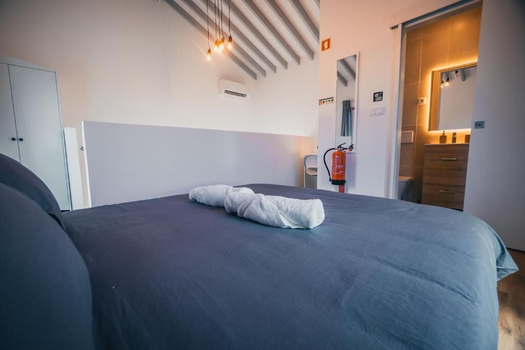A neatly made bed with a folded towel, a fire extinguisher, a wardrobe, and a mirror in a modern room.