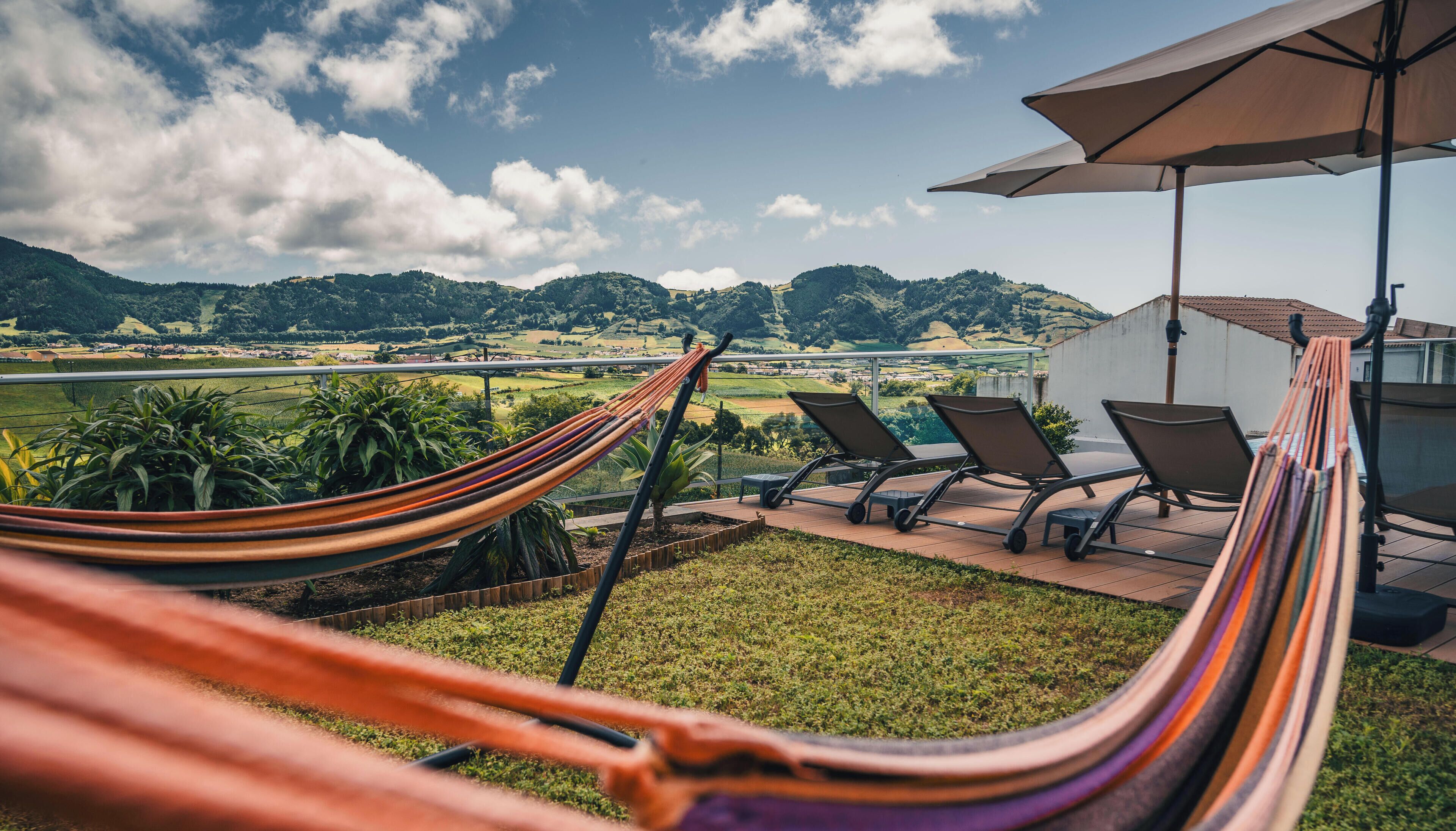 A hammock, lounge chairs, and an umbrella on a terrace with a view of a green landscape and hills.