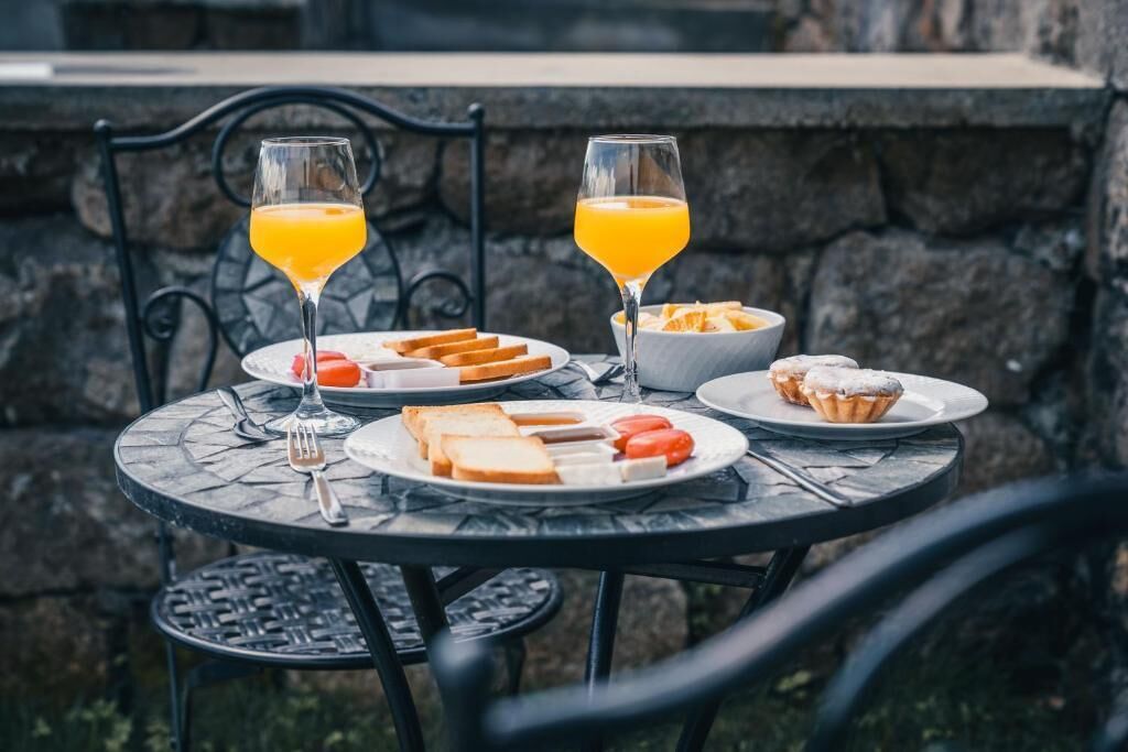 A round outdoor table with a stone wall backdrop, set with plates of toast, cheese, tomatoes, and a bowl of fruit. Two glasses of orange juice are also present.