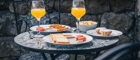 A round outdoor table with a stone wall backdrop, set with plates of toast, cheese, tomatoes, and a bowl of fruit. Two glasses of orange juice are also present.