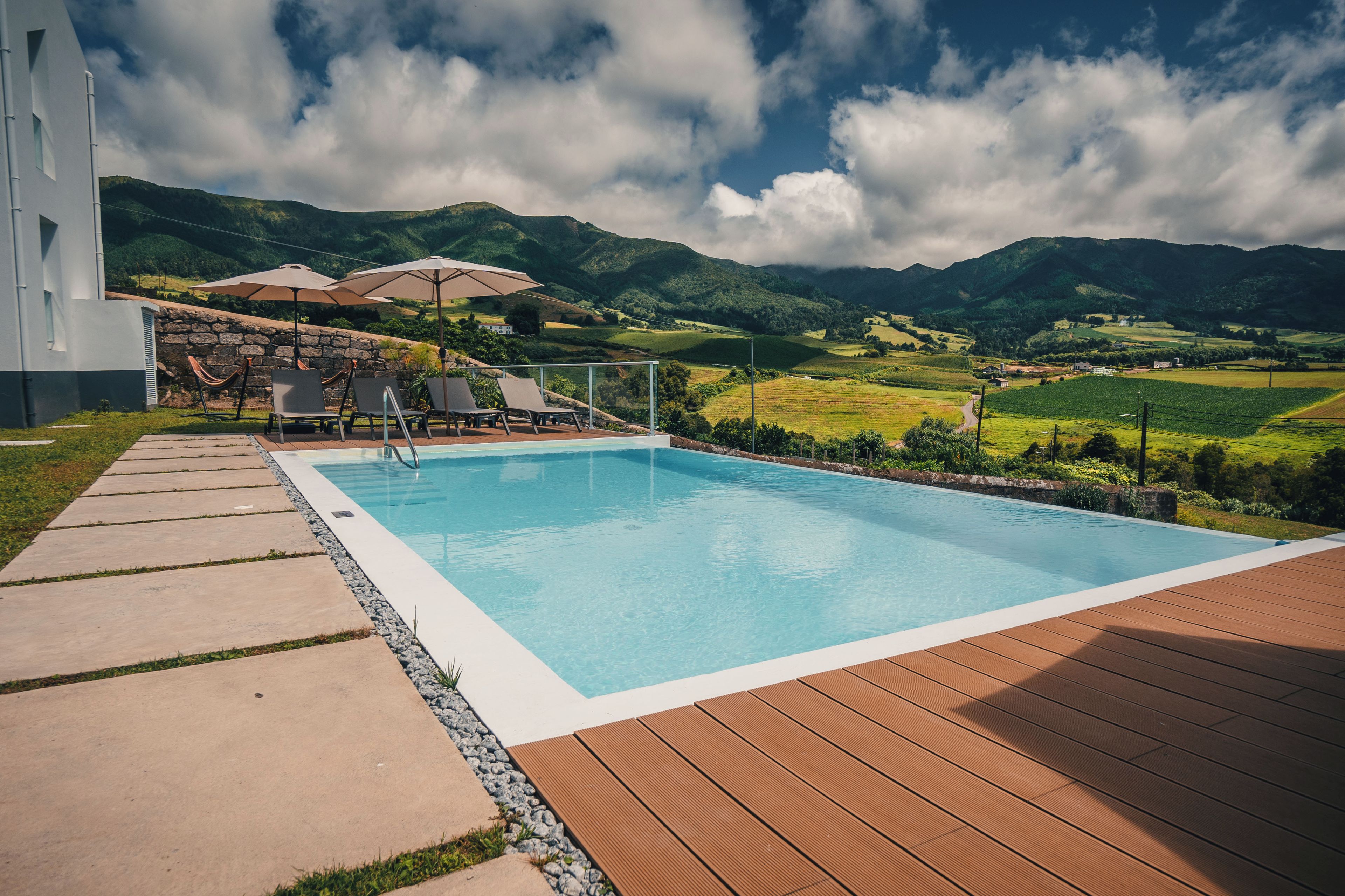 A rectangular swimming pool with a wooden deck in the foreground, surrounded by a stone wall and lounge chairs.