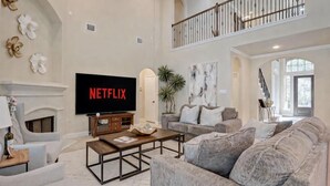 Uma sala de estar ampla com lareira, uma grande televisão de tela plana exibindo 'NETFLIX' e uma área de estar confortável com sofás e uma mesa de centro.