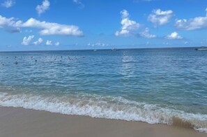 Una playa con agua azul cristalina, olas suaves y algunos barcos a lo lejos.