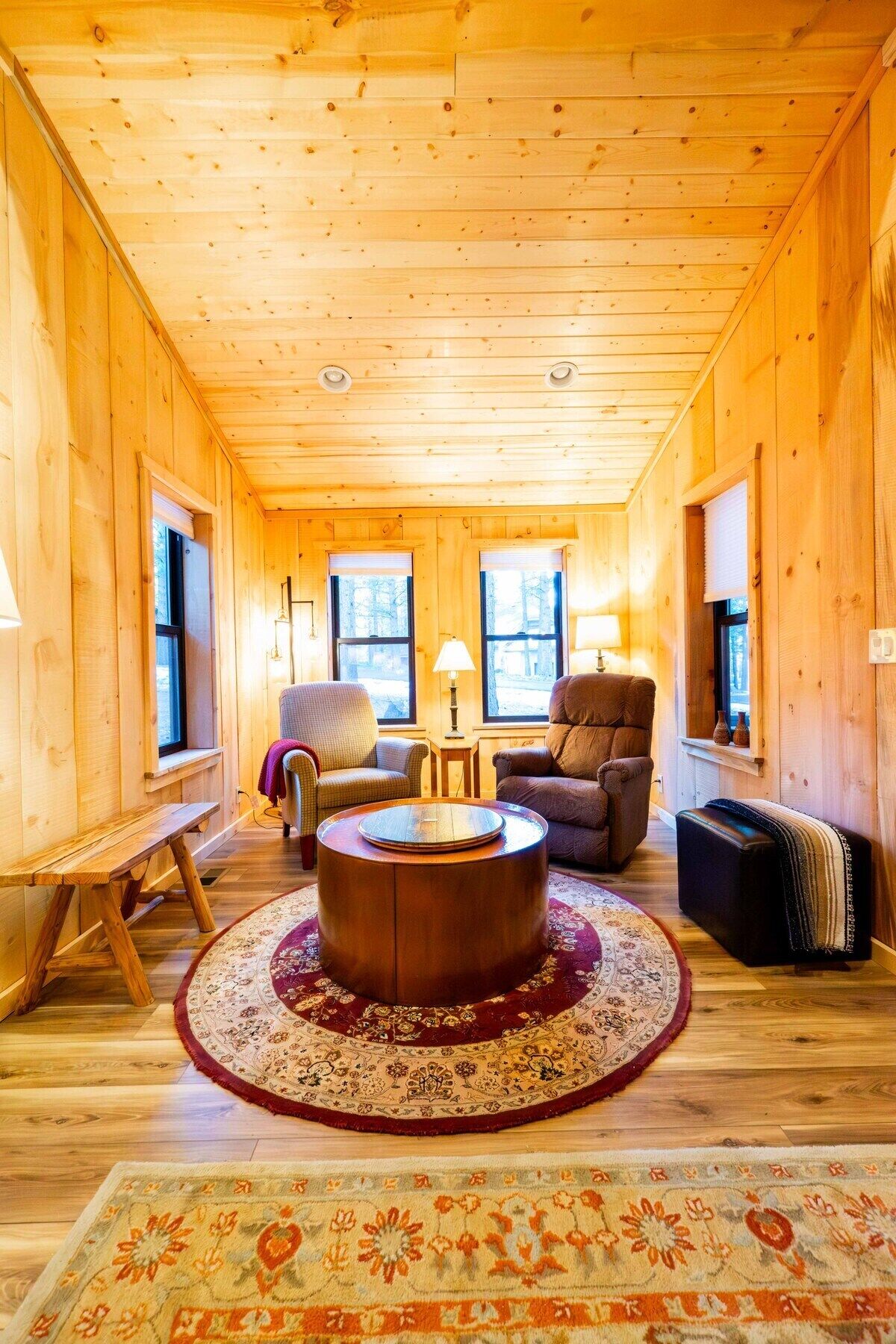 A cozy wooden cabin interior with a round coffee table, two armchairs, a bench, and a patterned rug.