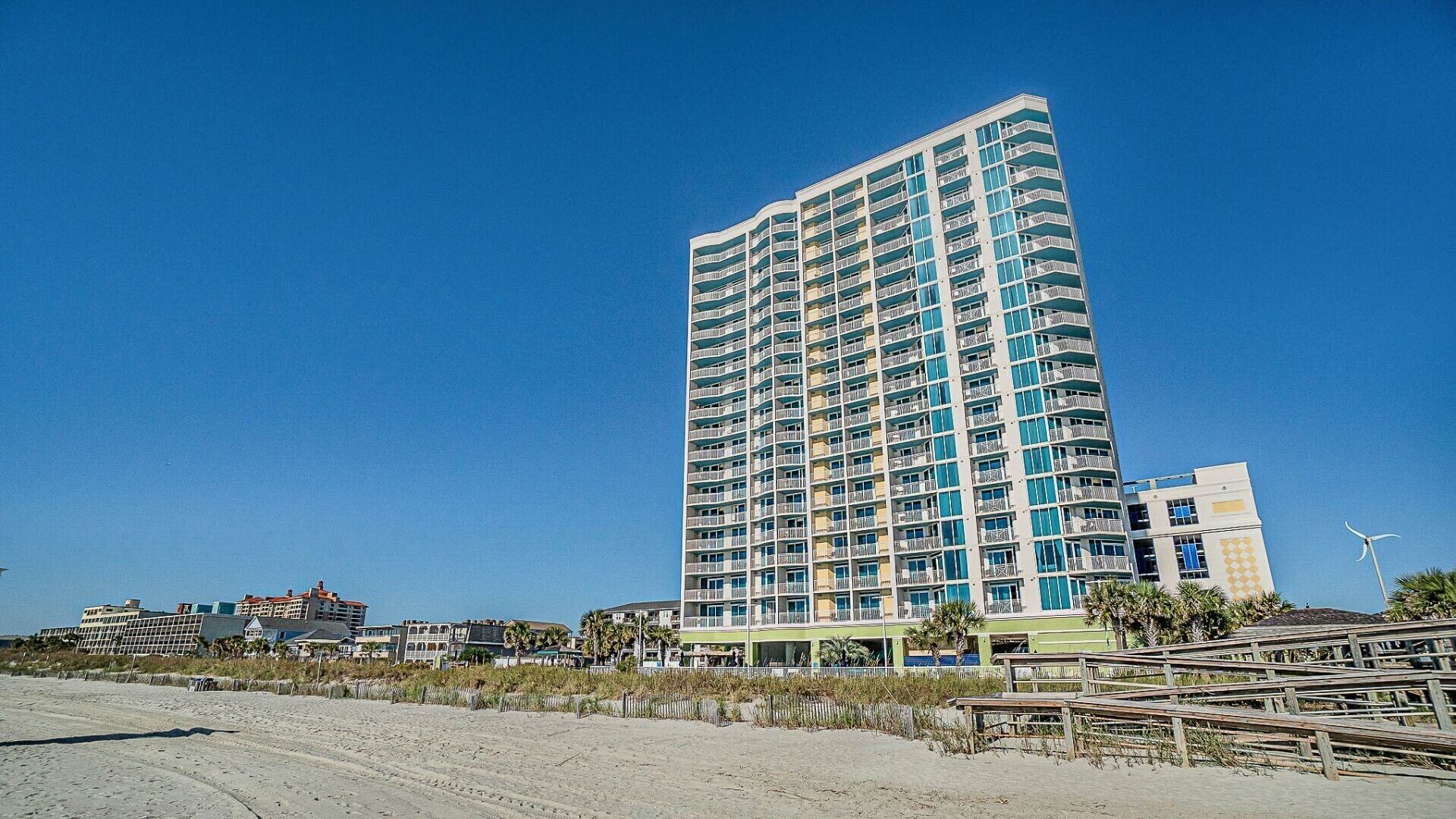 A tall, multi-story hotel with balconies, located near a beach with a sandy foreground and some vegetation.