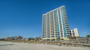 Een hoog, meerlaags hotel met balkons, nabij een strand met een zandvlakte en begroeiing.