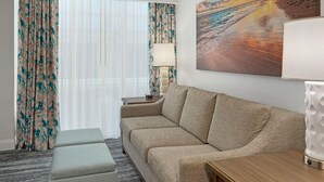 Hotelkamer met een beige bank, een klein voetenbankje, een houten bijzettafeltje en een lamp. Er is een groot raam met dunne gordijnen en een schilderij van een strandgezicht.
