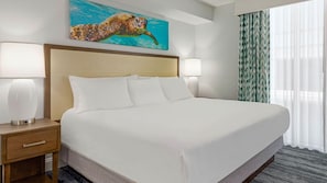 A hotel room with a large bed, two bedside lamps, a nightstand with a drawer, a painting of a sea turtle, and a window with sheer curtains. - 2 bdrm Myrtle Beach Resort (North Myrtle Beach)
