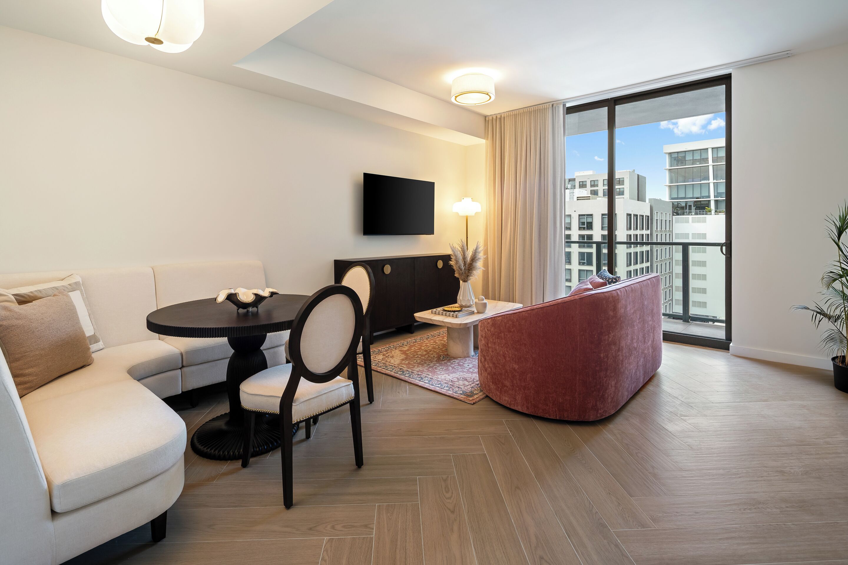 A modern living room with a sofa, dining table, chairs, a flat-screen TV, and a large window with a city view.