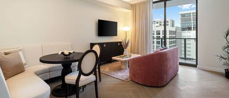 A modern living room with a sofa, dining table, chairs, a flat-screen TV, and a large window with a city view.