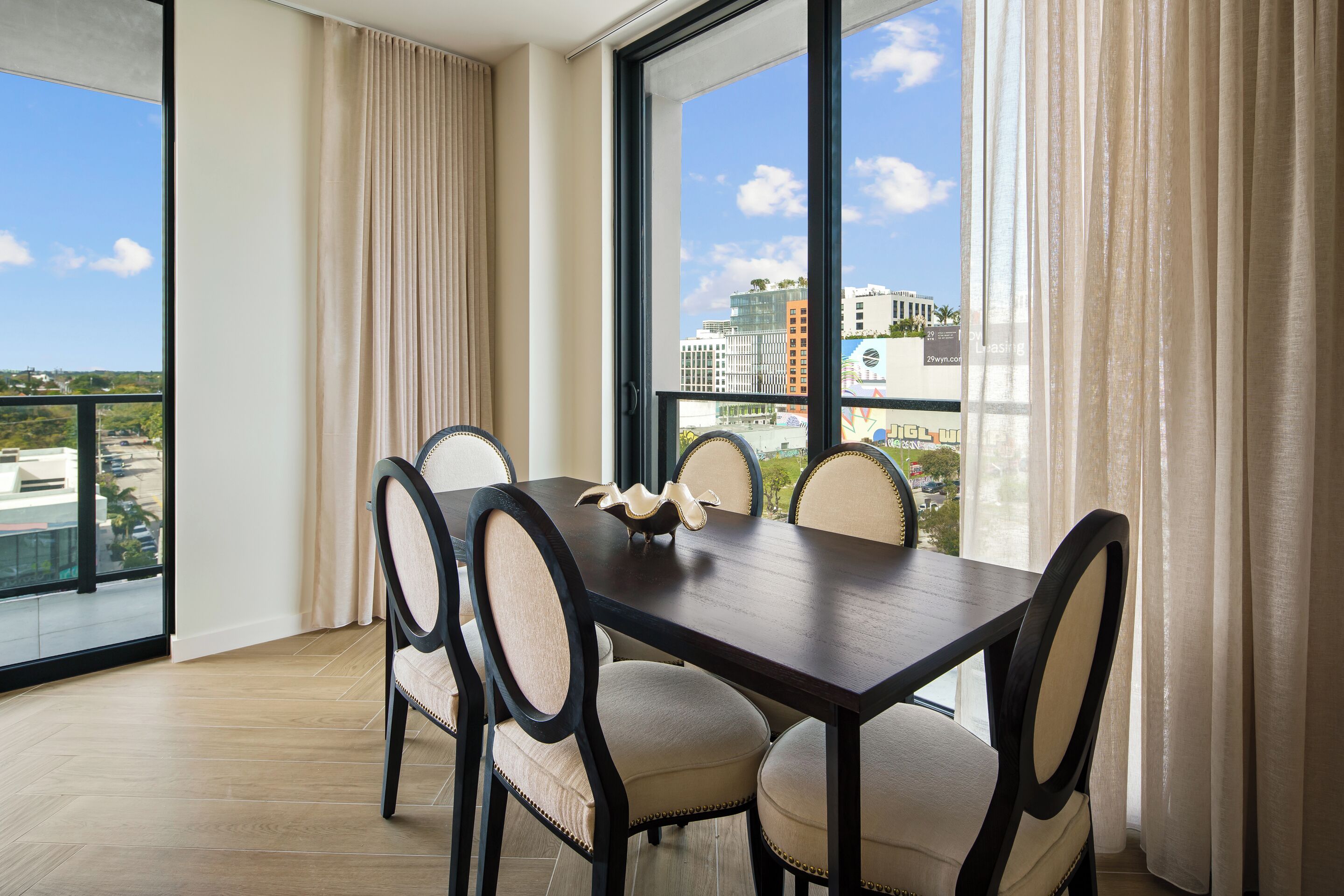 A dining area with a table and chairs, large windows with curtains, and a view of buildings.