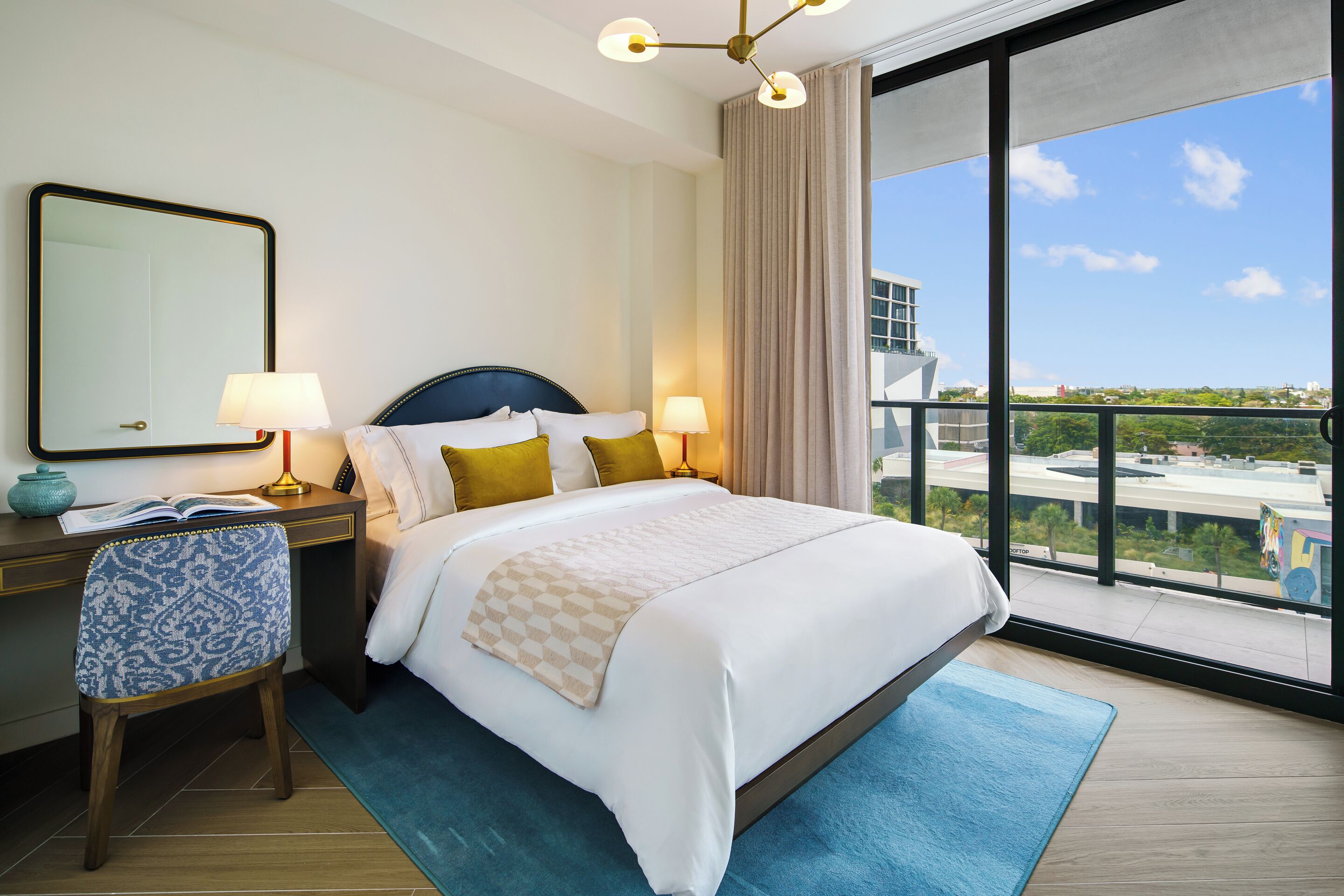 A hotel room with a large bed, two bedside lamps, a desk with a chair, a large window with a view, and a ceiling light fixture.