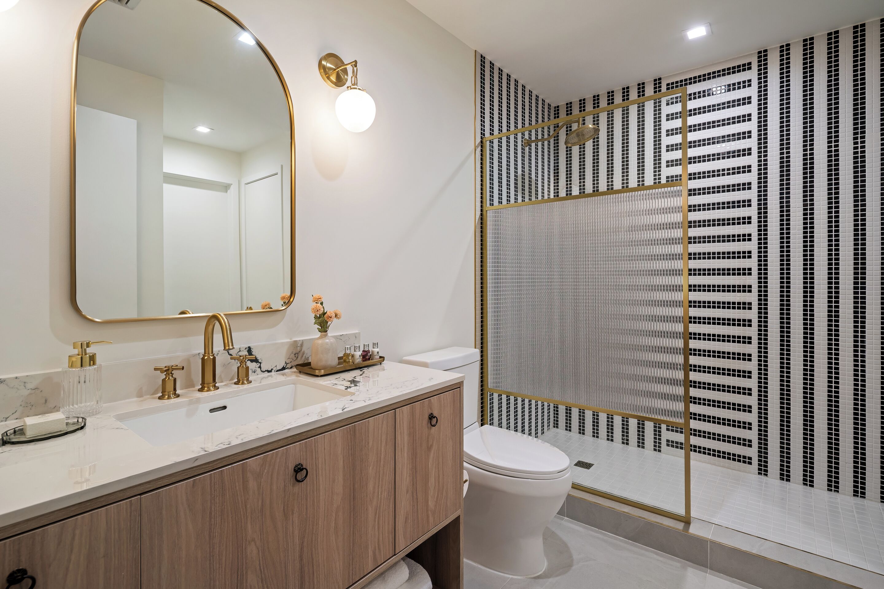Ein modernes Badezimmer mit einem großen Spiegel, goldenen Armaturen, einem weißen Waschbecken und einer Dusche mit einer Tür aus gemustertem Glas.