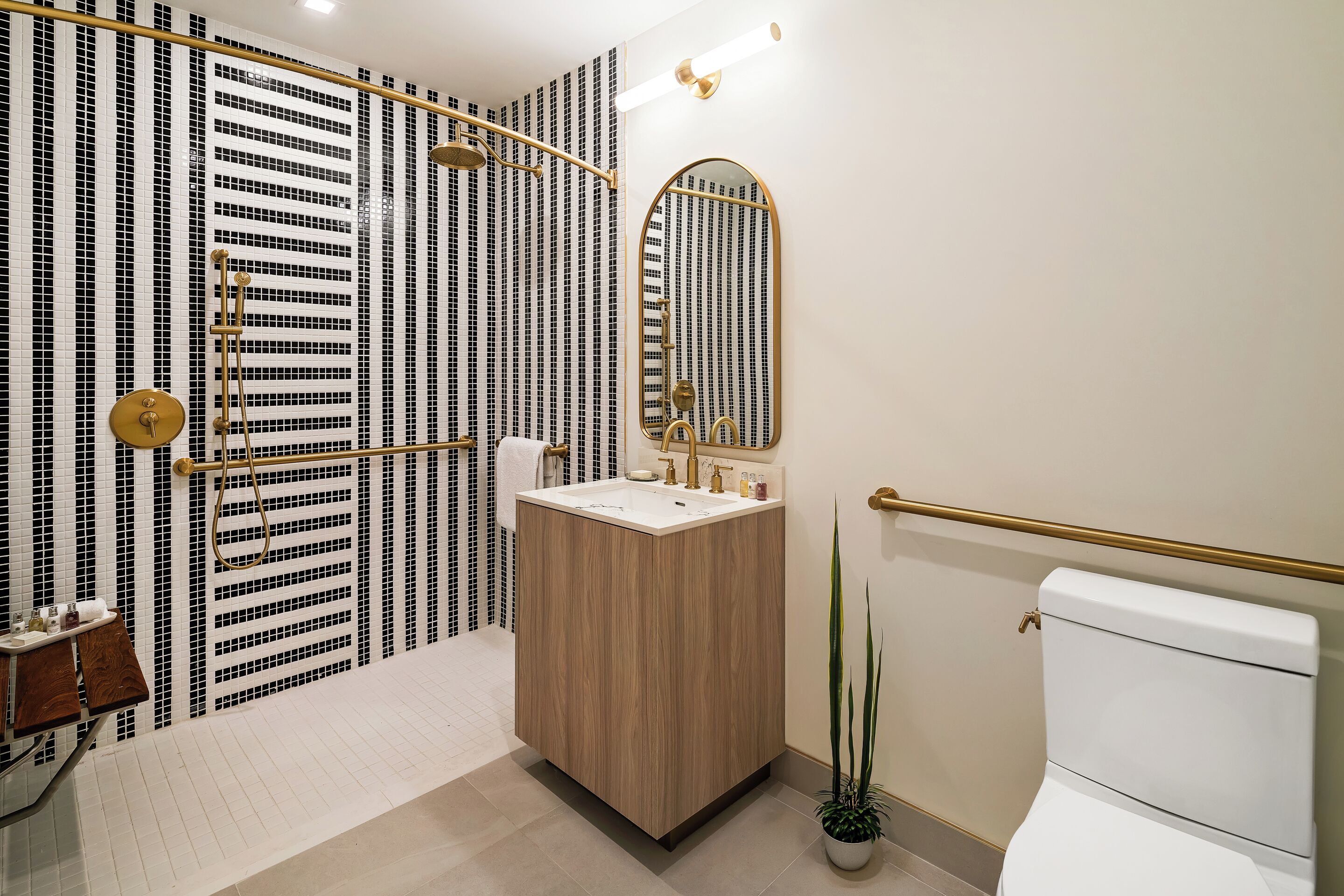 Un bagno moderno con uno specchio grande, una doccia con soffione dorato, un water e un lavandino con mobile contenitore in legno.