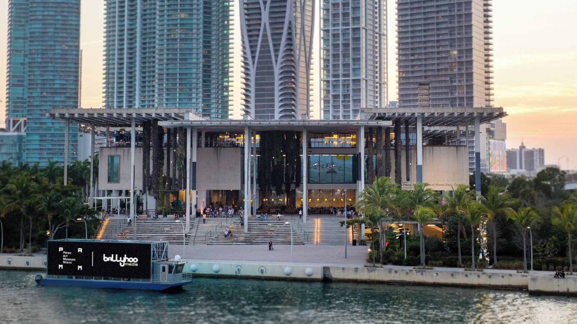 Un edificio moderno con una entrada amplia, un barco amarrado frente a él y altos rascacielos circundantes.