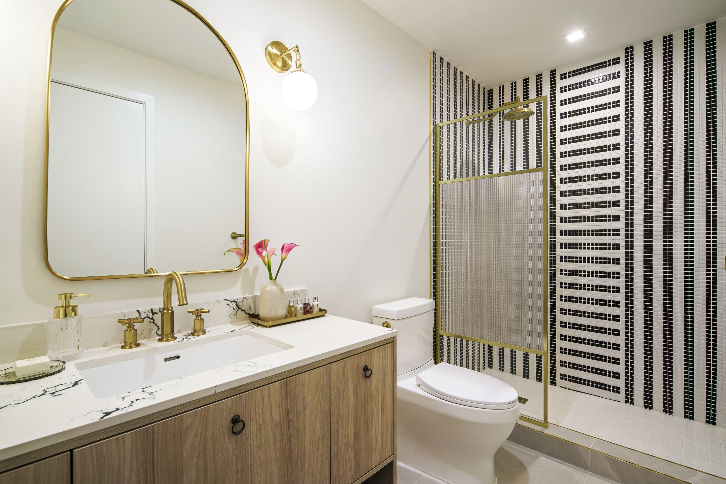 A bathroom with a white sink, gold faucets, a large mirror, and a shower with a black and white patterned wall.