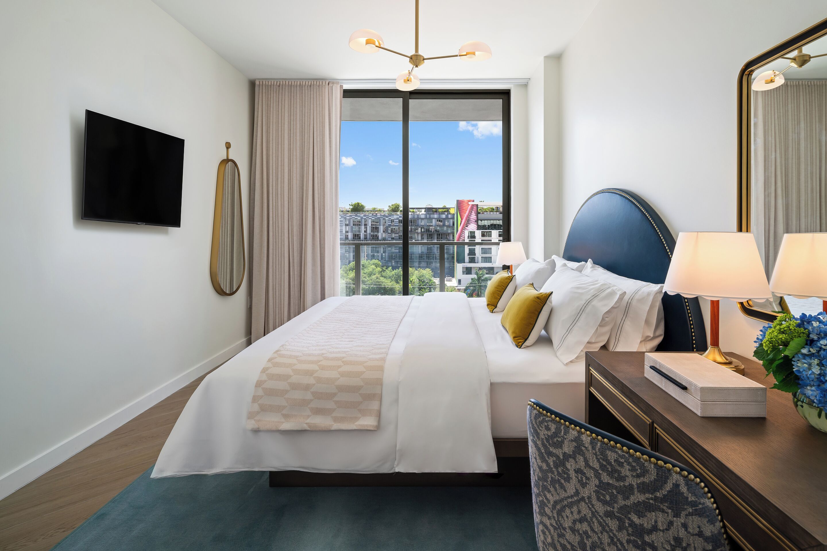 A hotel room with a large bed, a television, a desk with a lamp, and a view of buildings outside.