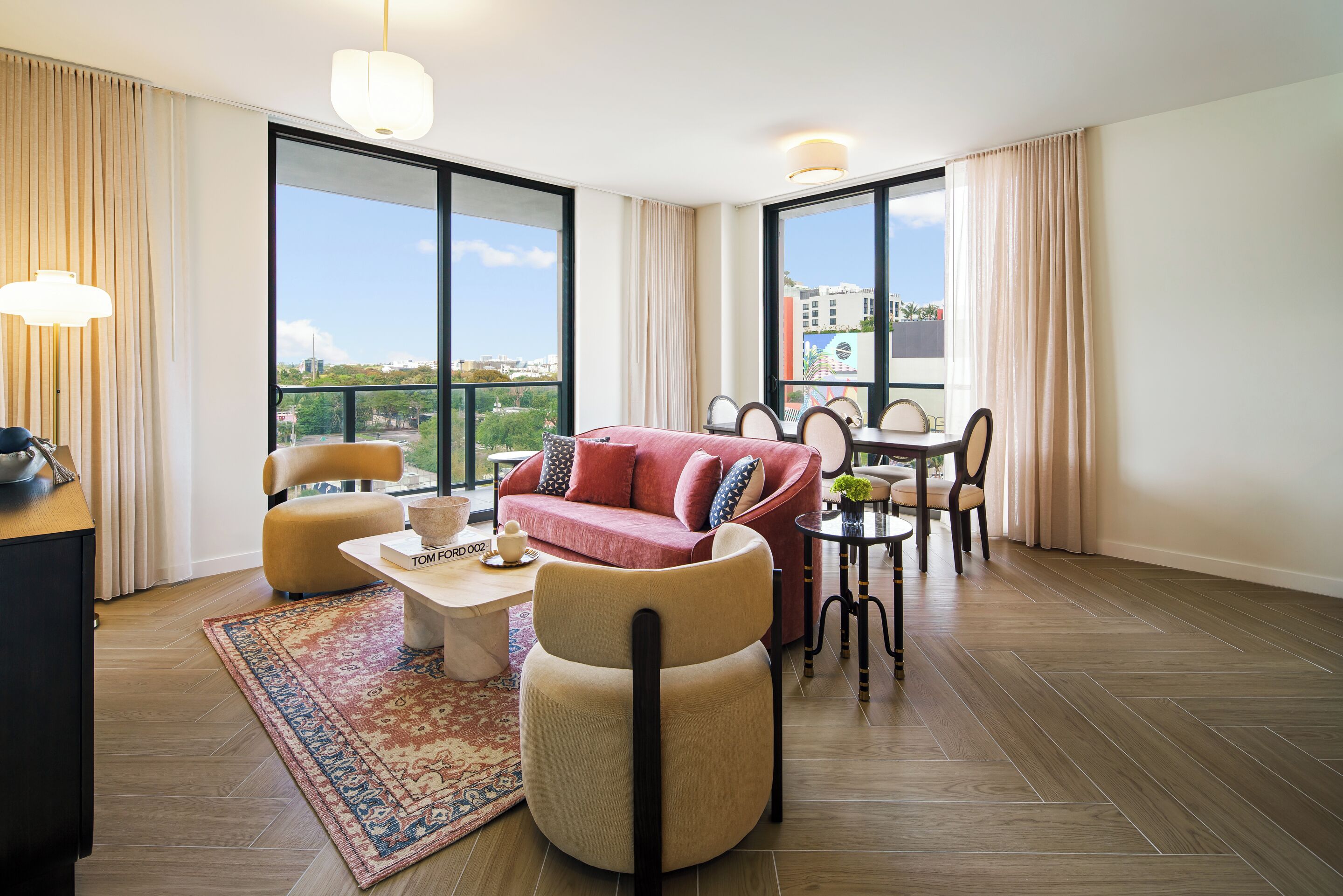 A modern living room with a sofa, chairs, a coffee table, and a dining area with a table and chairs. Large windows provide a view of the city.