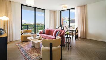 A modern living room with a sofa, chairs, a coffee table, and a dining area with a table and chairs. Large windows provide a view of the city.