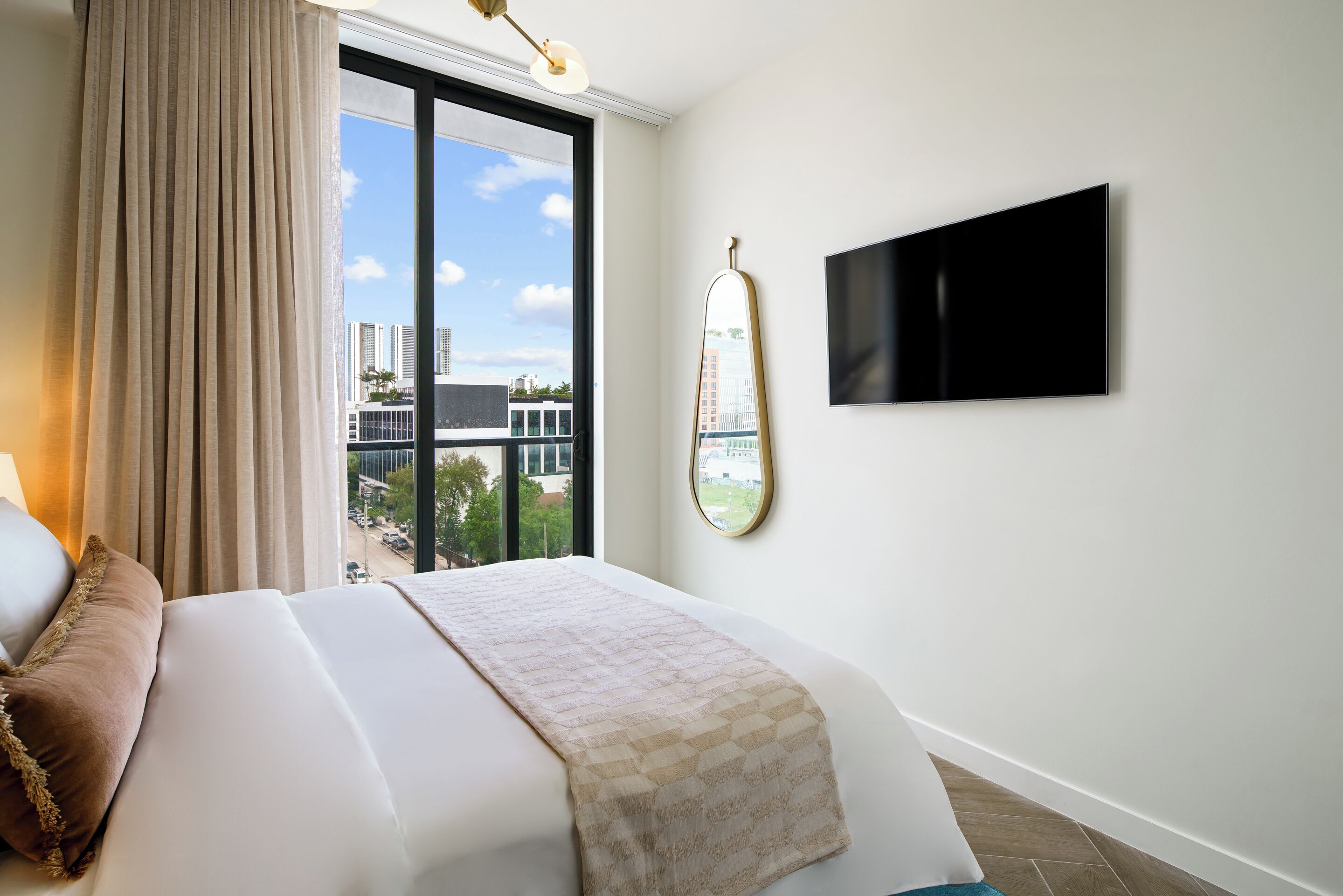 A hotel room with a large bed, a TV mounted on the wall, and a window with a view of buildings.