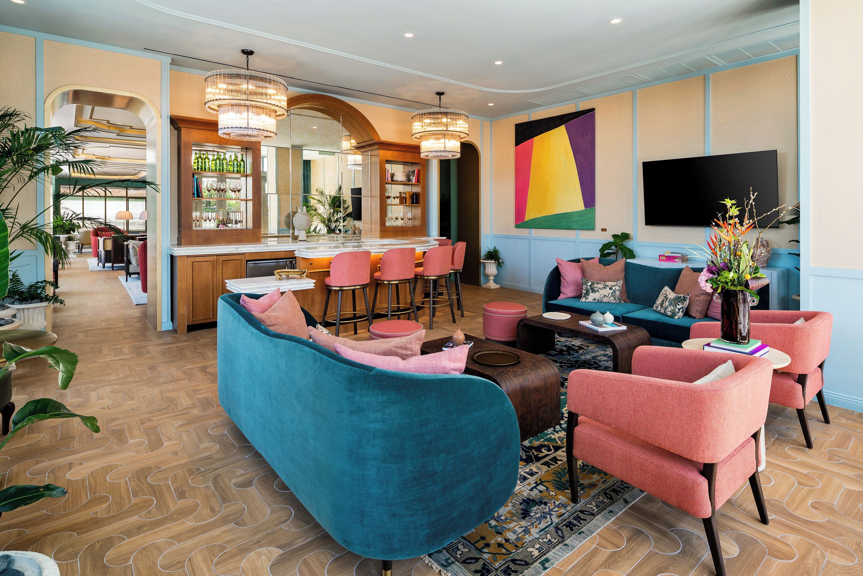 Un salon moderne avec un canapé turquoise, des fauteuils roses, un coin bar et un grand téléviseur.