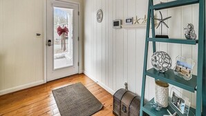A room with wooden flooring, a door with a red bow, a teal shelf with decorative items, and a small clock on the wall. - Classic 3BR House | Porch, Pool & Beach Nearby (Bethany Beach)