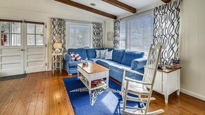 A living room with a blue sofa, wooden coffee table, white rocking chair, and patterned curtains. - Classic 3BR House | Porch, Pool & Beach Nearby (Bethany Beach)