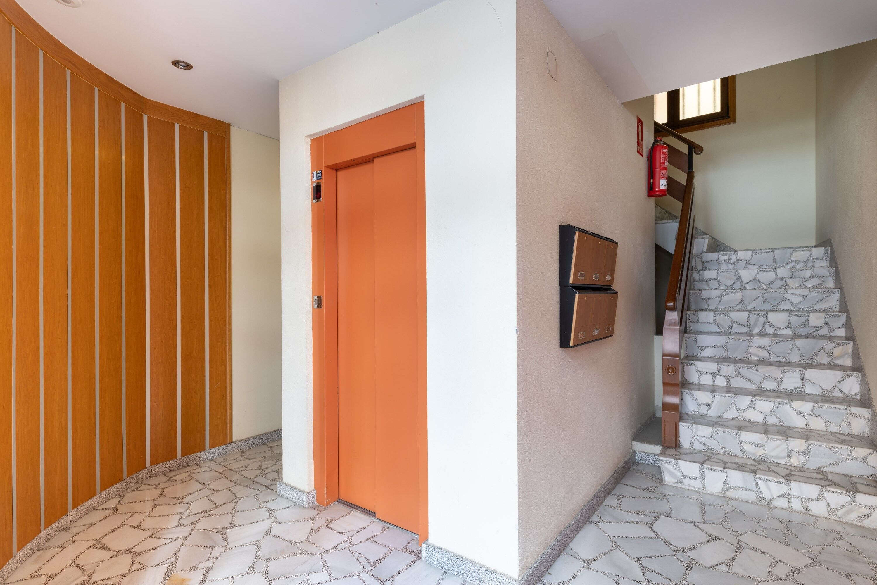 Un pasillo con panel de madera, una puerta anaranjada, una escalera con escalones de piedra y un extintor de incendios montado en la pared.