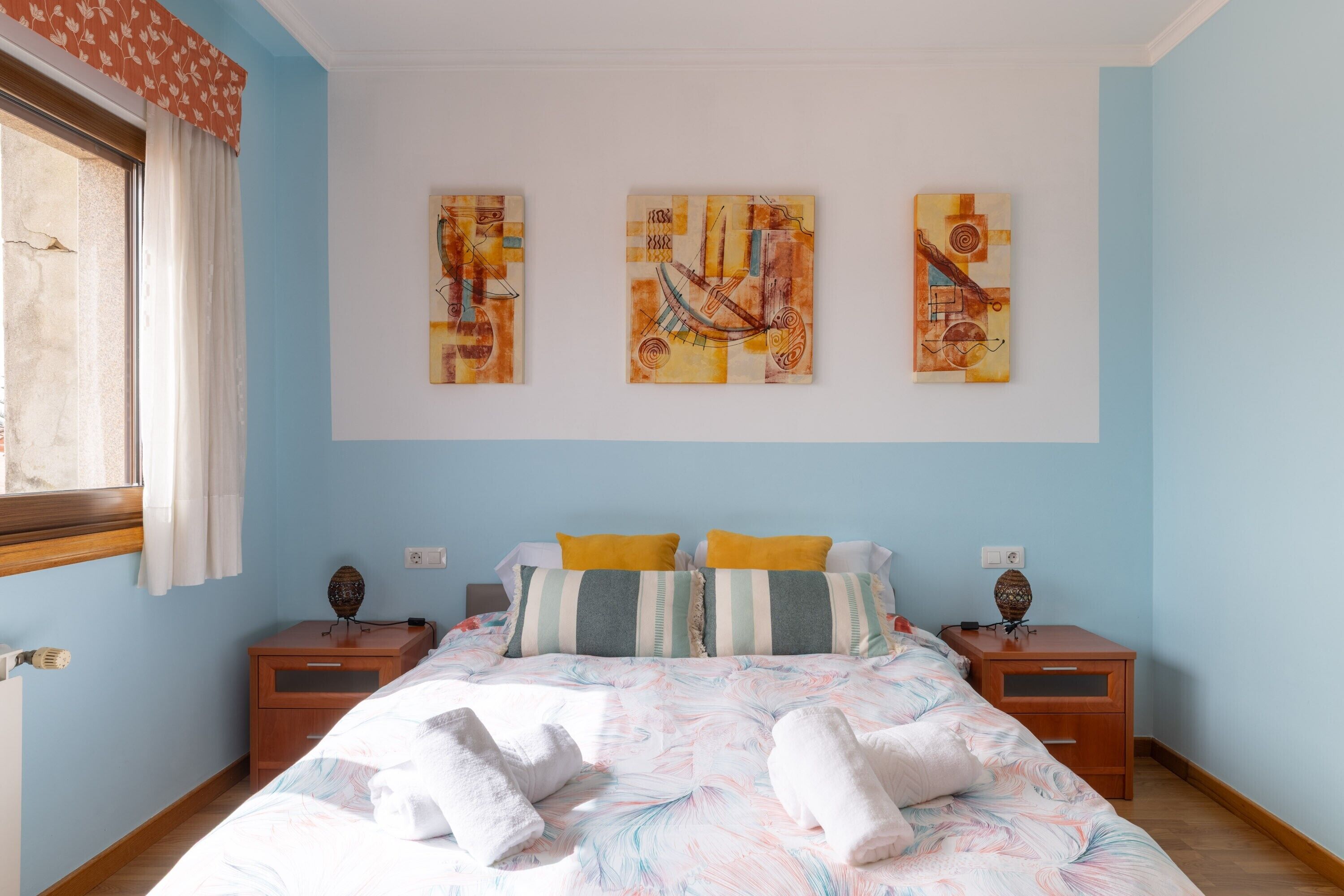 Een slaapkamer met een bed, twee nachtkastjes, een raam met gordijnen en drie kunstwerken aan de muur.