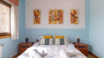 Een slaapkamer met een bed, twee nachtkastjes, een raam met gordijnen en drie kunstwerken aan de muur.
