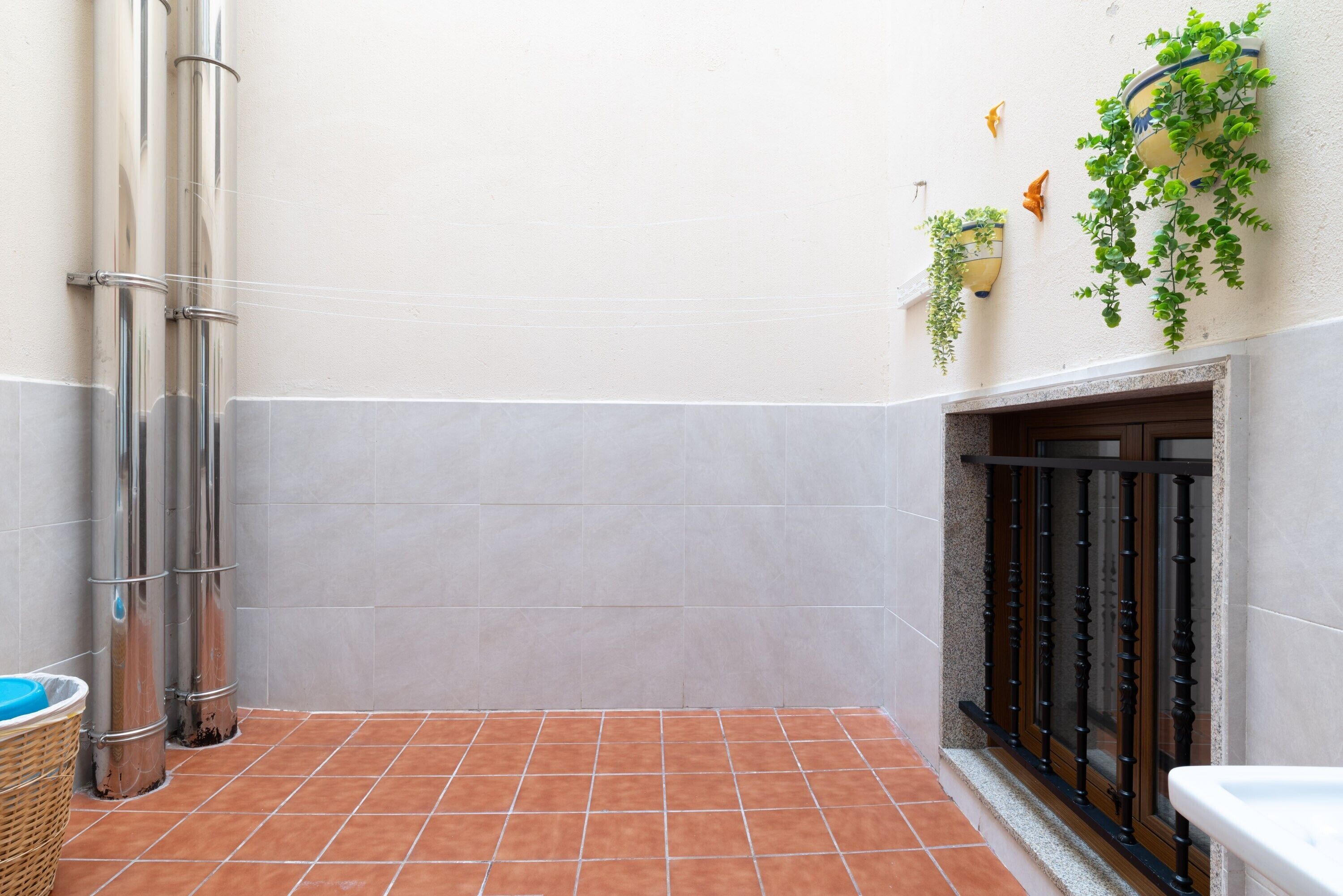 Eine geflieste Terrasse mit Metallkonstruktion, einem an der Wand befestigten Pflanzgefäß mit Grünpflanzen und einem Fenster mit schwarzem Metallgitter.