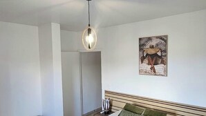 Een slaapkamer met een groot bed, een houten nachtkastje, een klein houten kistje, een kunstwerk aan de muur en een plafondlamp.