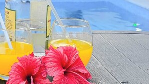 Een glazen fles en twee glaasjes met een gele vloeistof, een rode bloem en een zwembad op de achtergrond.