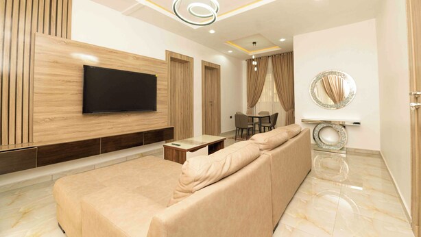 Ein modernes Wohnzimmer mit einem großen Flachbildfernseher, einem beige gefärbten Sofa, einem Essbereich mit Tisch und Stühlen sowie einem dekorativen Spiegel an der Wand.