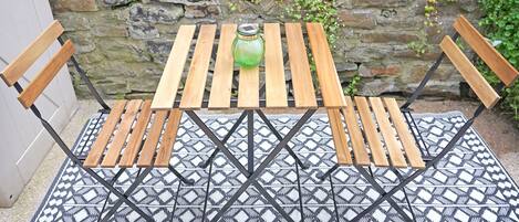 A wooden table and chairs set on a patterned rug, with a stone wall and greenery in the background.
