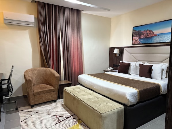 A hotel room with a bed, a chair, a desk, an air conditioner, and a painting of a coastal scene. - Vinpy Hotels (Uyo)