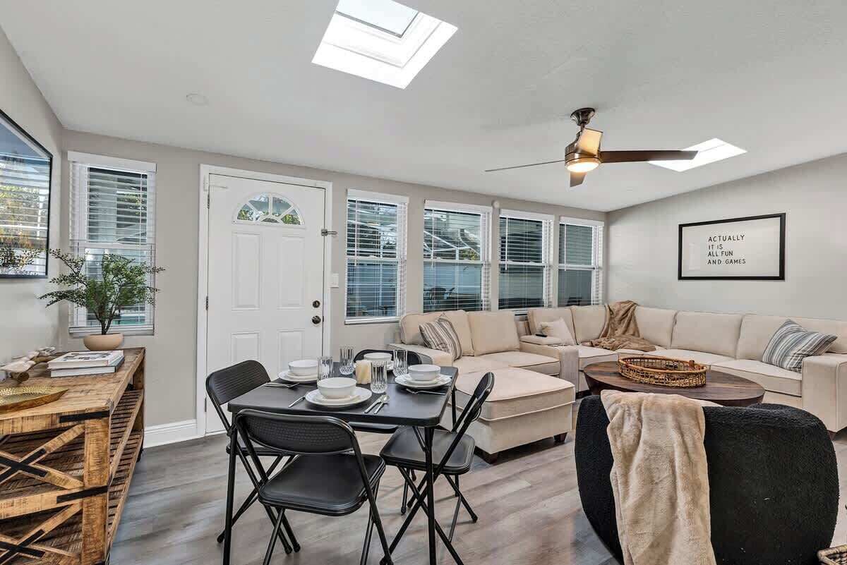 A modern living room with a dining area, a ceiling fan, and a large window with blinds.