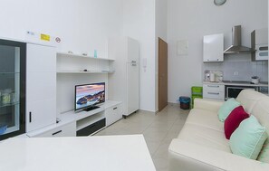 Un salon moderne doté d’un téléviseur à écran plat, de meubles blancs, d’un canapé avec des coussins et d’un coin cuisine avec des placards et un four à micro-ondes.