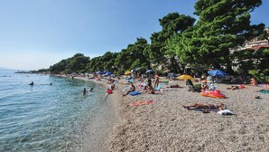 A pebbled beach with people sunbathing and swimming, surrounded by trees and umbrellas. - Gorgeous apartment in Brela with WiFi (Brela)