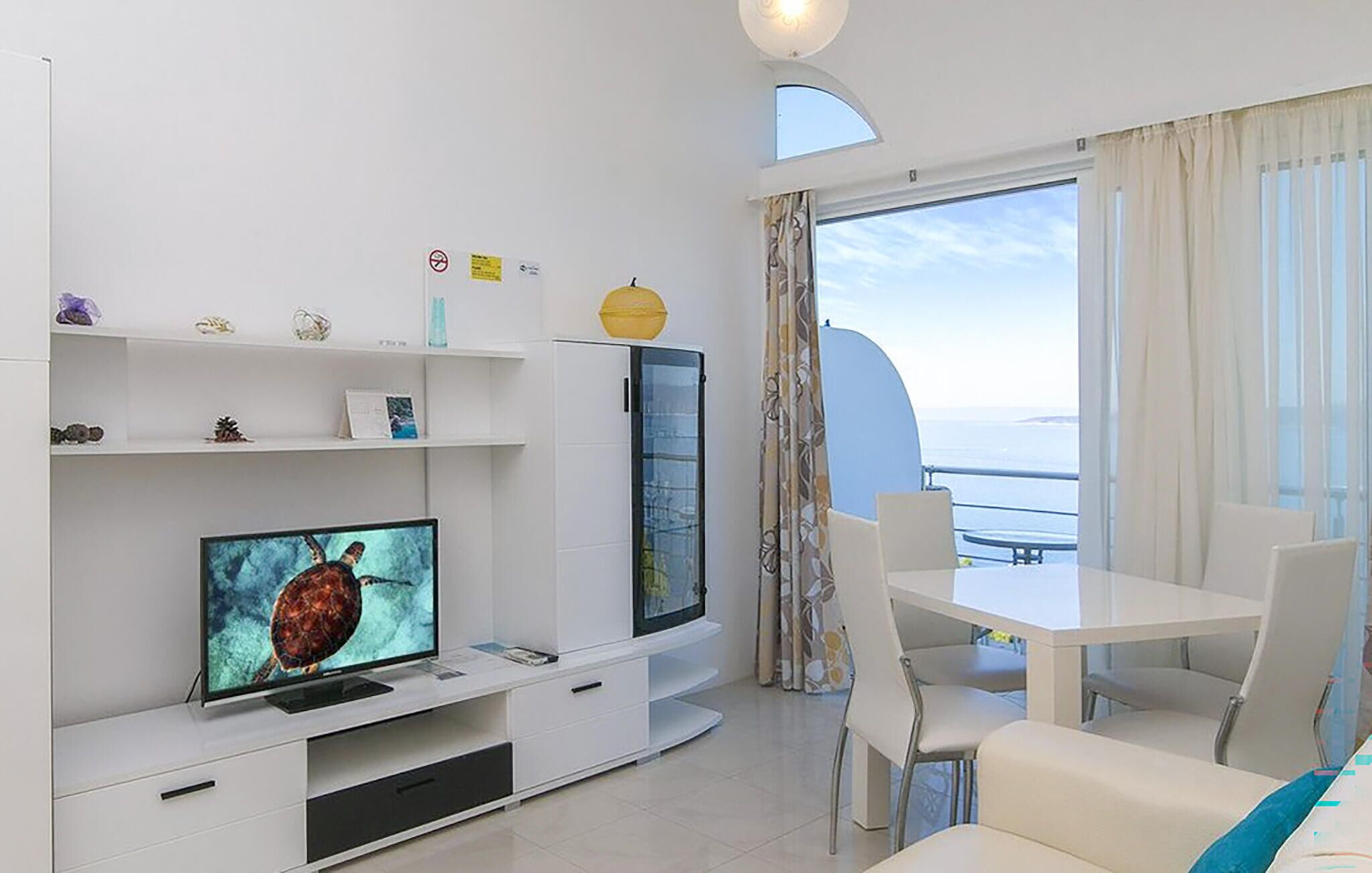 Una sala de estar moderna con un televisor de pantalla plana, un comedor con mesa y sillas, y un ventanal con vistas al mar.
